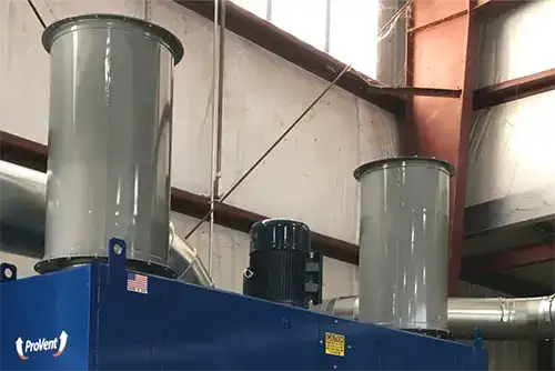 Top section of a blue ProVent industrial dust collection system with two large gray exhaust ducts and a motor unit, located inside an industrial facility with visible metal beams and overhead piping.