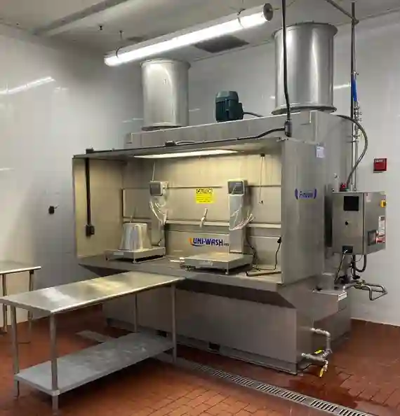 Stainless steel UNI-WASH industrial cleaning station with overhead exhaust vents, two workstations, and a control panel, set up in a clean, tile-floored room. The system is equipped with lighting, ventilation ducts, and adjacent stainless steel work tables for efficient industrial washing and filtration processes.