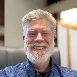 Headshot of Paul, a man with a full white beard and short hair, smiling in an indoor office environment.