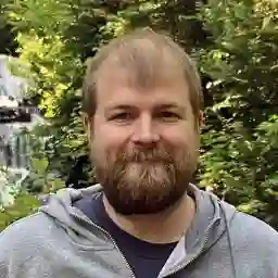 Portrait of Kevin, a man with a beard, smiling in an outdoor setting with lush greenery and a waterfall in the background.