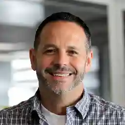 Headshot of Dave, a man with short dark hair and a friendly smile, wearing a plaid shirt in an indoor setting.