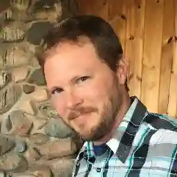 Portrait of Andrew, a man with short brown hair and a beard, wearing a plaid shirt in front of a stone wall backdrop.